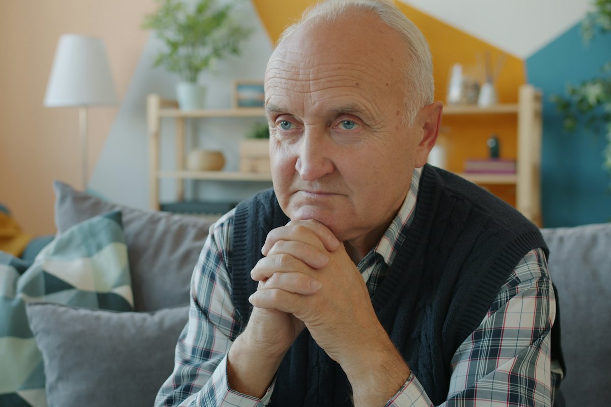 A retired man sitting on his couch with his hands clasped, looking thoughtful